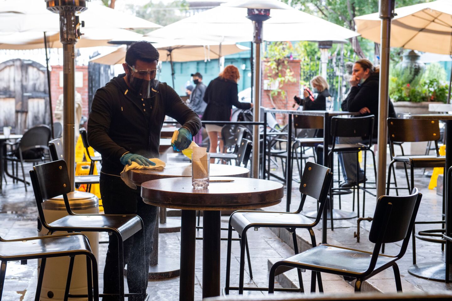 A male employee wearing a face mask, face shield and gloves wipes down an outdoor bar table under heat lamps and umbrellas at The Abbey in West Hollywood.