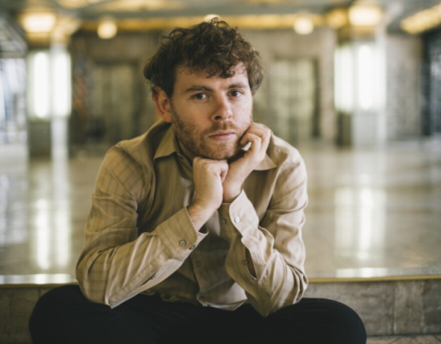 Gabriel sits cross legged on a shiny floor, hands under chin, with brown curly hair, wearing a beige striped shirt