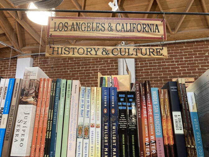 A row of paperback books on a shelf with a brick wall in the background. Two small wooden signs hanging above the books read "Los Angeles & California" and "History & Culture."