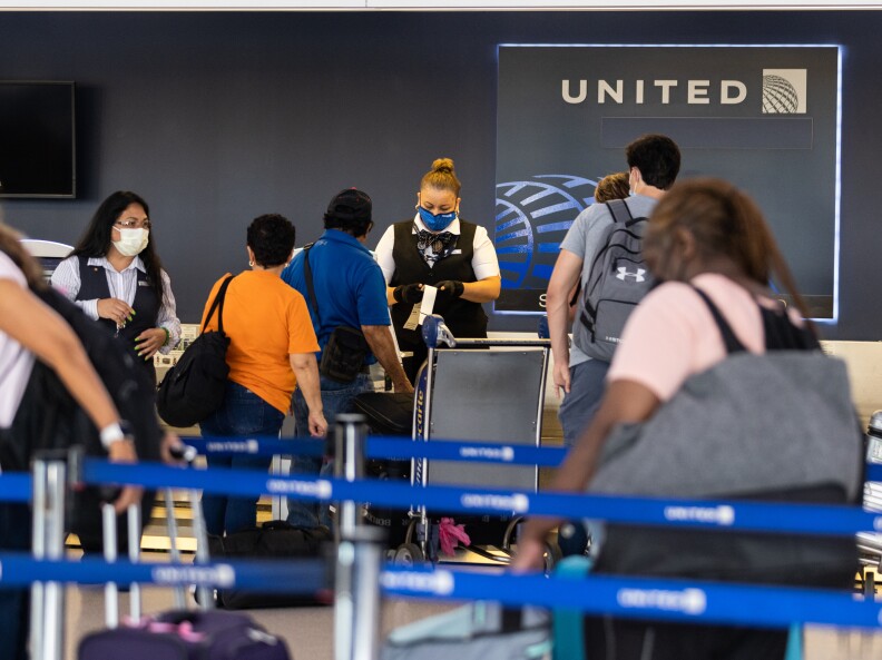 A line of people wait between blue ropes in front of a terminal with the words "UNITED AIRLINES." The woman checking them in wears a United uniform and a mask. Most travelers are dressed for warm weather and carry shoulder bags.