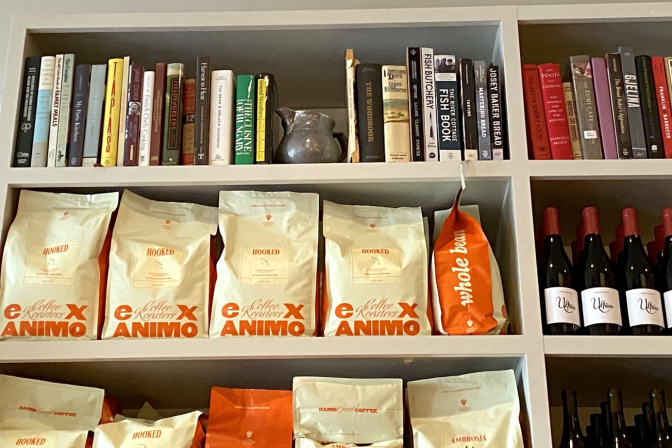 A bookcase has a series of cookbooks on the top shelf, with cream and orange coffee bags and bottles of wine below.