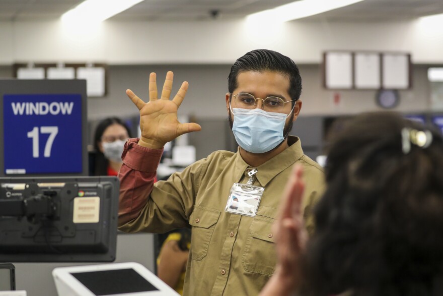 A man holds up his hands with his five fingers outstretched. He wears a mask and a tan shirt, with a red undershirt. He is in front of Window 17.