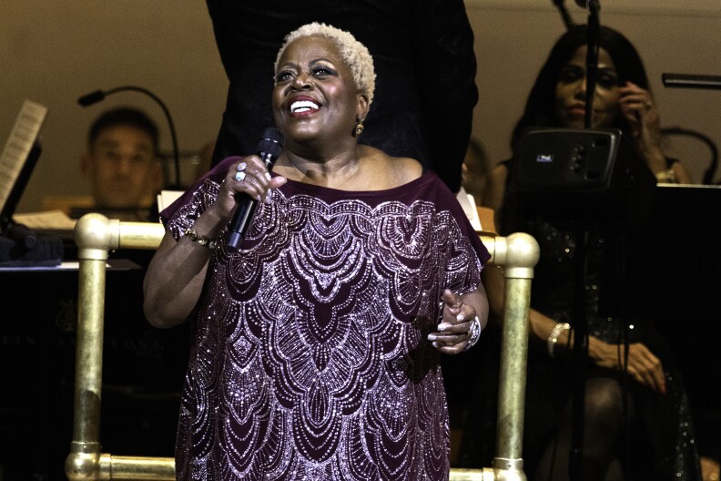 Lillias White smiles mid-performance while holding a microphone. She's a Black woman with short, platinum blonde hair, wearing a sparkling purple dress.