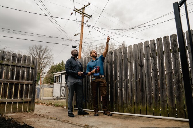 Real estate appraiser Jack Sonceau (left) and his trainee, Devin Minnis, assess a house in Baltimore. Devin raises his left arm towards something out of frame. The two are flanked by two sets of ashy brown colored fences. They stand under a cloudy grey sky, with power lines intertwining above them. In the background are brain trees and a small peak of a white house.