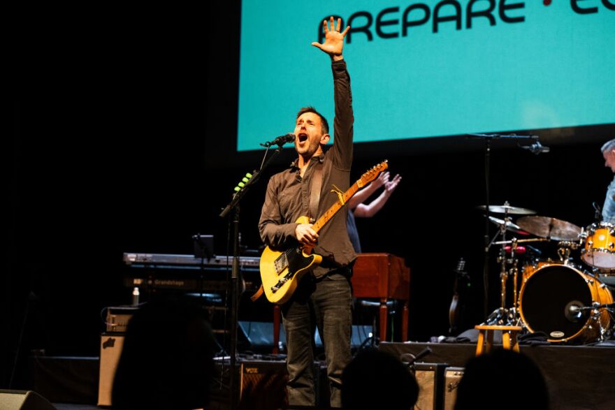 Man wearing a strapped guitar, dark jeans and a dark button up shirt has his left arm stretched out above him as he sings into a microphone on stage with a drummer, pianist and screen behind him.
