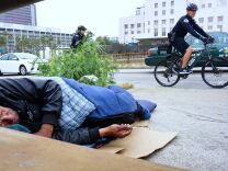 Los Angeles Police Department officers patrol on bicycles past a homeless man napping with cigarette in hand at his encampment on a downtown sidewalk in Los Angeles, California on June 7, 2017, a city facing a growing homeless population and less affordable housing.    / AFP PHOTO / FREDERIC J. BROWN        (Photo credit should read FREDERIC J. BROWN/AFP/Getty Images)