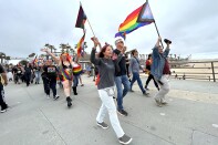 A crowd of peope march on the pier with their hands in the air with pride flags. Many people have rainbow colors on their clothing in some way.