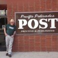 A man in a tan hat, a dark green shirt, gray pants and brown shoes leans against a brick wall with a sign that says "Pacific Palisades Post Printing & Publishing". The man smiles at the camera. A glass door behind him has the address "839" on it.