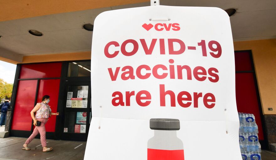 A woman walks toward the doors of a CVS Pharmacy in the background as a large sign advertises in big red text that COVID-19 vaccines are available at the location.