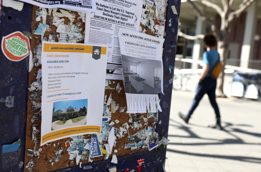 A bulletin board with ripped up remnants of old listings, as well as multiple flyers for student housing with photos of the homes, inside and outside. In the background, a student walks by, blurred focus.