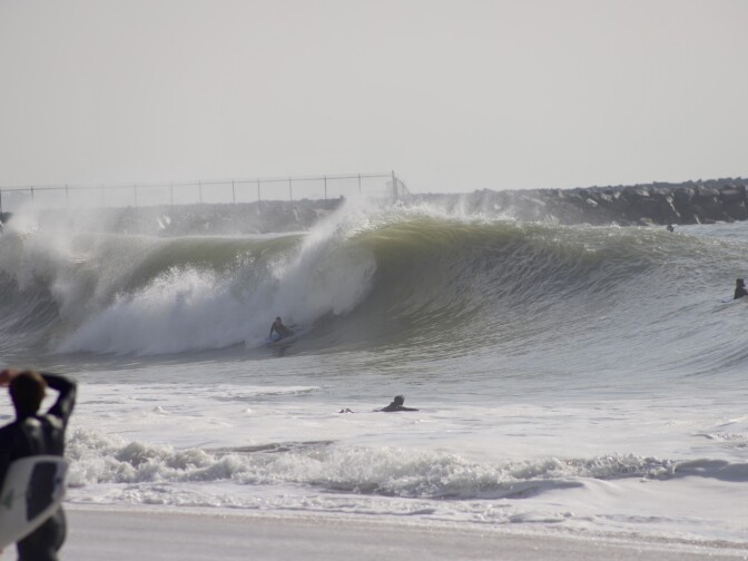 A body boarder taking a big wave in front of a jetty. A surfer looks on from shore. 
