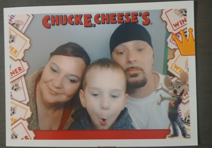 A photograph with a red Chuck E. Cheese's logo across the top and a cartoon mouse in the bottom corner. Two adults and a young child are framed in the photo.