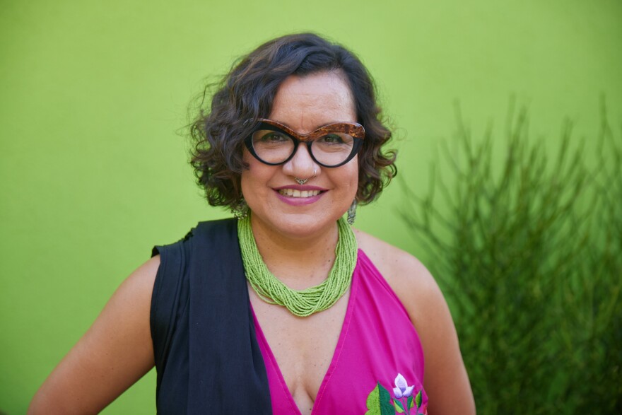 Musician and professor Martha Gonzalez wears horn rimmed glasses and fuchsia top and stands in front of solid green wall and plant.