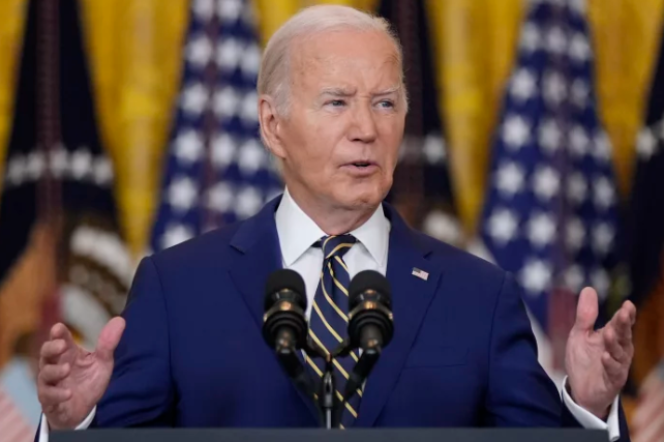 A man with white hair is wearing a blue suit jacket, white shirt, and a striped tie with an American flag pin. He has both hands open and is speaking behind a podium behind a mic with flags in the background. 