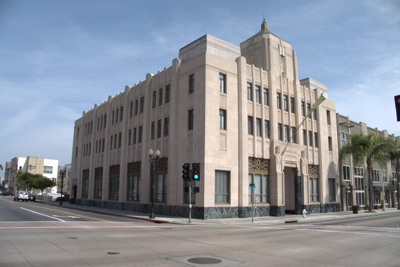 A tan rectangular art deco building, three stories, sits at the corner of an intersection of two gray roads in front of a blue sky. An inscription on the right side of the building reads "Santa Ana City Hall."