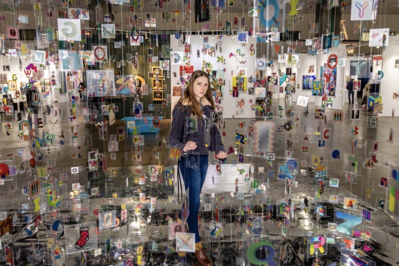 A woman stands in the middle of an art piece made of small square paintings hung from the ceiling by strings.