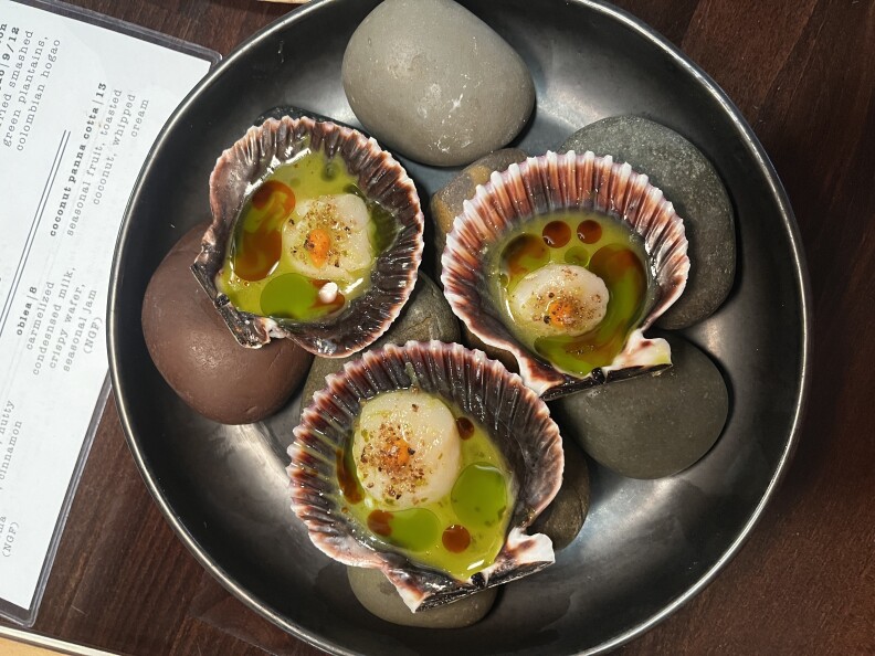 Three scallops are served on a half shell which rests in a shallow black bowl filled with grey river rocks. The shells contain petite scallops which are topped with a small amount of orange pepper sauce and a sprinkling of red powder. They are swimming in a liquid bath of different shades of green and red. The visible outer rim of the inside of the shell has a dark purple color.