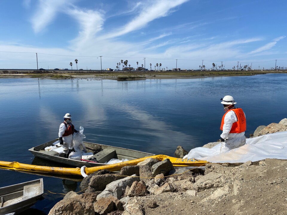 A worker on shore wearings a bright orange life preserver, a white hard hat and respirator as he works with another person in a row boat wearing a dark life preserver and similar head gear. Both are in white coveralls. A yellow flexible tube runs from the water onshore.