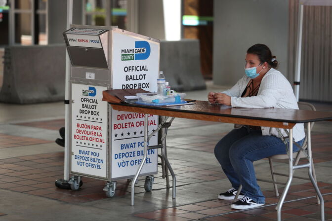 MIAMI, FLORIDA - AUGUST 11: A poll worker waits for voters to help them put their mail-in ballot in an official Miami-Dade County ballot drop box on August 11, 2020 in Miami, Florida.  The secure drop boxes at polling stations allow voters to drop off the state’s Primary Election Vote-By-Mail ballots and skip any voting lines as people try to vote and stay healthy during the coronavirus pandemic. The ballot drop box is also a way to securely deliver a voting ballot to the polling station and not have to mail the Vote-By-Mail ballot through the U.S. Postal Service. (Photo by Joe Raedle/Getty Images)