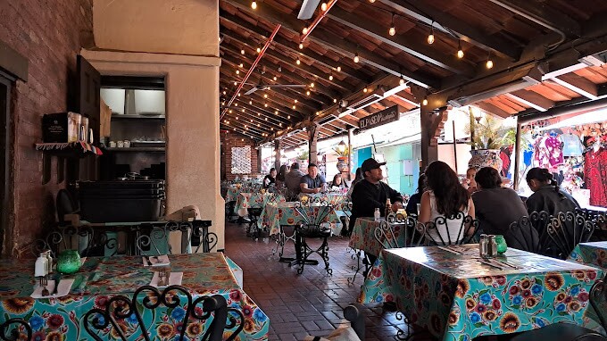 Covered patio at El Paseo Inn in the heart of Olivera Street with string lights, floral tablecloths, and wrought-iron chairs, where diners gather over meals in a lively, colorful setting.