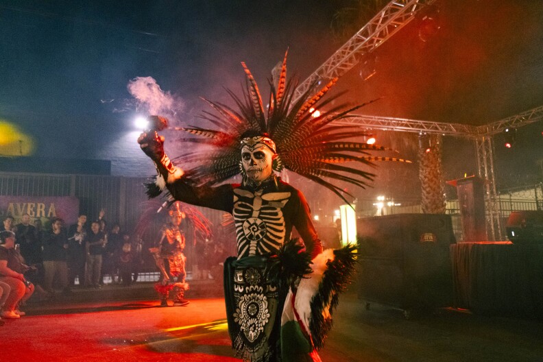 A wide shot of a man covered in skeleton face paint and clothing while holding up a cup of copal, a burning incese. He's wearing a large feathered headress and other cultural materials to do the dance.