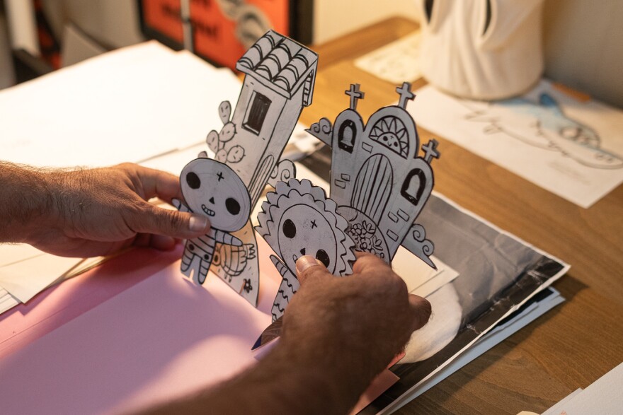 Paper dolls of a boy and girl skeleton along with a cut-out church and building with a shingle roof held up by a man's hands.