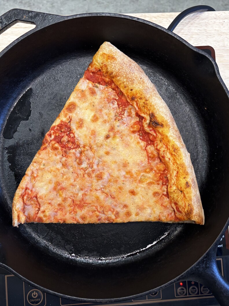 A single slice of cheese pizza sits inside of round black cast iron pan on a light wood surface.