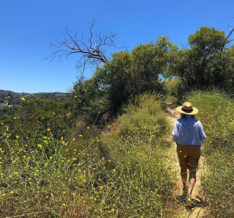 A person in shorts, blue shirt and a sun hat hikes on a dirt trail