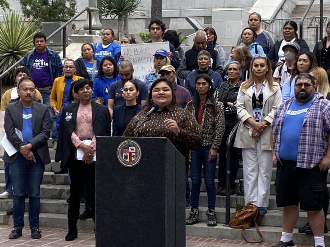 LA City Council member Eunisses Hernandez speaks at a podium that has the seal of Los Angeles on it. There is a crowd of more than a dozen behind her. Some wear blue t-shirts that mark them as supporters of the progressive policy advocacy group LA Forward. 