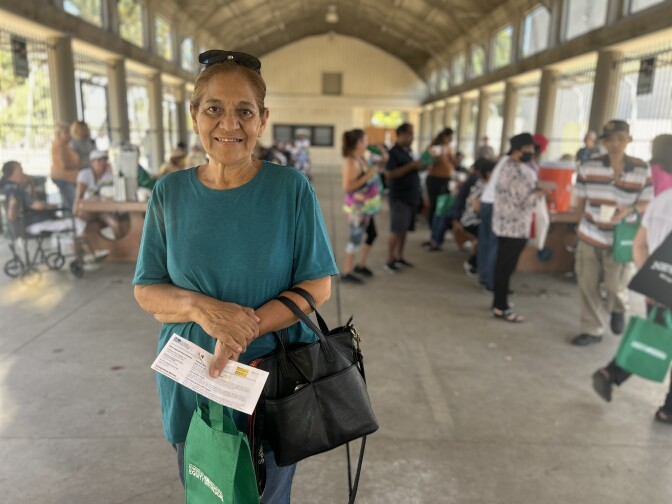 An older woman with light brown skin wears a teal short-sleeved shirt and holds a bag and folded paper and smiles for the camera with sunglasses pushed onto her head. 