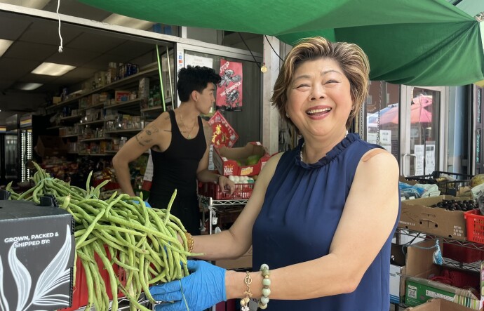 An Asian woman stands at the entrance of a small market under a green tarp, smiling and holding a bunch of long green beans. Behind her, a younger Asian man stands amid stacked produce boxes, and shelves of groceries line the back.