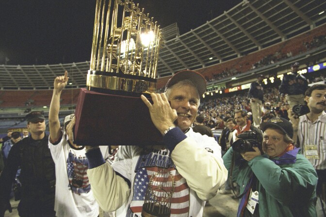 A man wearing a baseball cap and a white shirt with the American flag on it hoists a large trophy on his right shoulder. A group of people stand behind him. They are outside, in a baseball stadium.