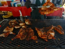 A vendor grills and flips seasoned chicken over an open flame at an outdoor food stand, with various supplies and drinks in the background.