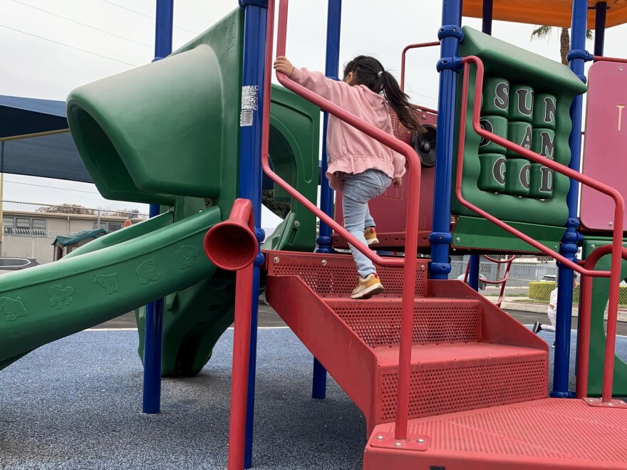 A small girl in a pink jacket, light blue jeans and yellow sneakers climbs up the red stairs of a jungle gym.
