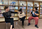 An 8th grader wearing a black t-shirt, reddish pants and black shoes sits in a chair close to two adult women. The women watch the 8th grader's hands are mid-motion in front of his face as he communicates in American Sign Language.