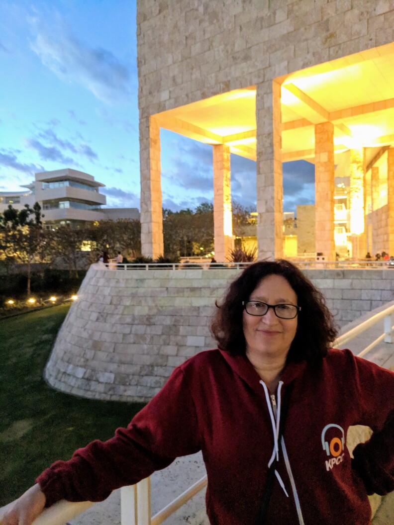 A woman with red hair and glasses stands outside of the Getty museum at dusk. She has one arm up on a railing and is wearing a maroon colored KPCC hoodie.