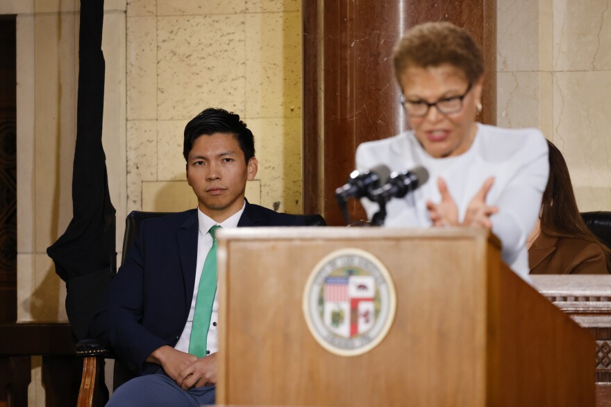 An Asian man is sitting in a chair on the left side of the photo, over the shoulder of an out-of-focus Black woman speaking into a microphone on a wooden podium. The man is wearing a mint green tie and black suit.