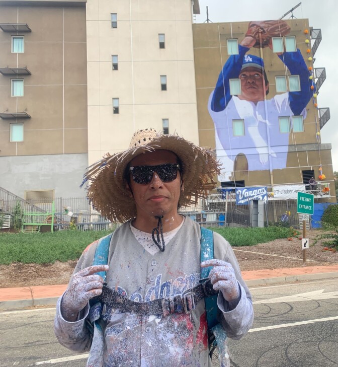 A Latino man wearing a grey baseball jersey, a straw hat, black sunglasses covered in paint stands in front of an apartment building with the mural of another Latino man posed as catching a ball. 