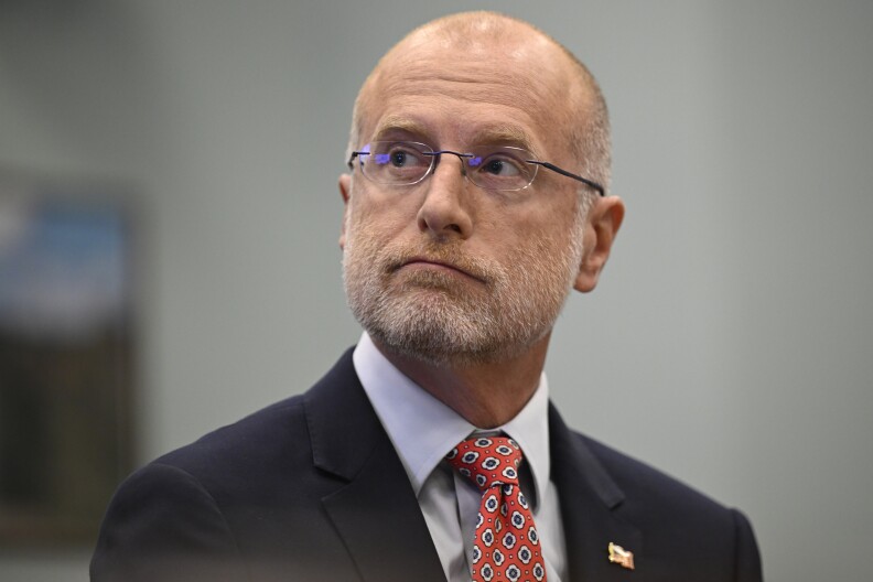 A white man with gray facial hair wears rimless glasses and a suit.