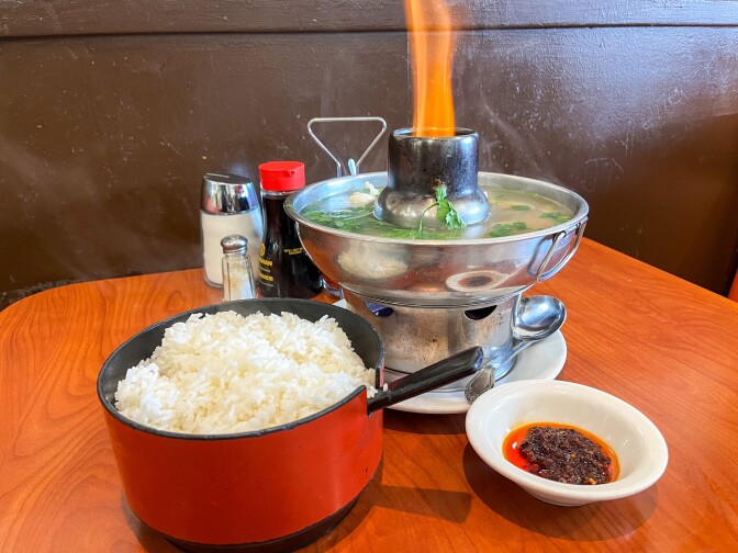 A collection of dishes is arranged on a medium brown, rounded tabletop against a dark brown-painted wall. In the center is a large metal hot pot filled with soup, featuring a raised middle section and an orange flame. The pot's base is filled with a clear liquid infused with green herbs. In front of the soup, a round red container holds steamed white rice, and to the right, there is a small white dish containing red chili paste.
