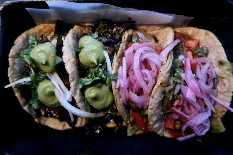 four tacos on corn tortillas. two are topped with dabs of green sauce. the other two are topped with pickled onions 