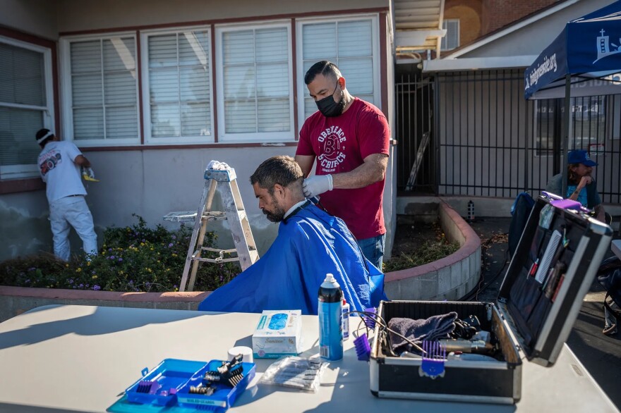 A light brown-skinned man in a red T-shirt, wearing a mask and gloves, gives a haircut to a light-skinned man sitting down wearing a blue haircut smock. Two other people are in the background, as well as a building. Equipment is on a table in front.