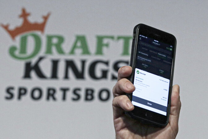 A medium skin toned hand holds up a phone with a prediction market app open, while the DraftKings logo can be seen in the background on a white wall.