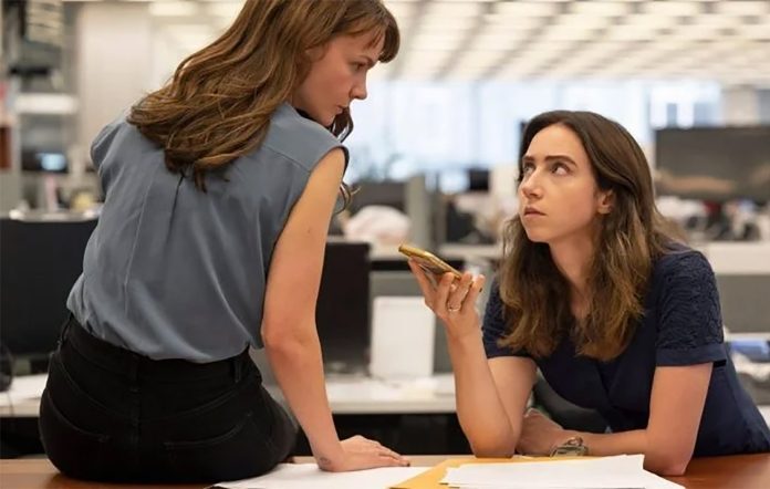 Carey Mulligan sits left of the frame on top of a long wood table. Her back is towards the viewer as she leans slightly to her right looking across her shoulder at Zoe Kazan who sits right of her. Mulligan's face can be seen in profile, her expression serious and tense. She wears a grey tank tucked into black pants, her right arm braced on the table to help her lean. Zoe Kazan leans over the table to Mulligan's right. Kazan faces the viewers as she looks at Mulligan, her body angled left. Her left arm rests in front of her on the edge of the table while her right arm is propped up on her elbow as she holds a gold colored phone in her hand. It is facing up as if on speaker phone with someone. Kazan wears a navy blue t-shirt. She looks up at Mulligan only with her eyes, she expression is also tense. They women seem to be sharing a knowing look with each other as they listen to whatever is being said on the phone. Behind them are empty rows of black desktop computers and white walls of a newsroom. 