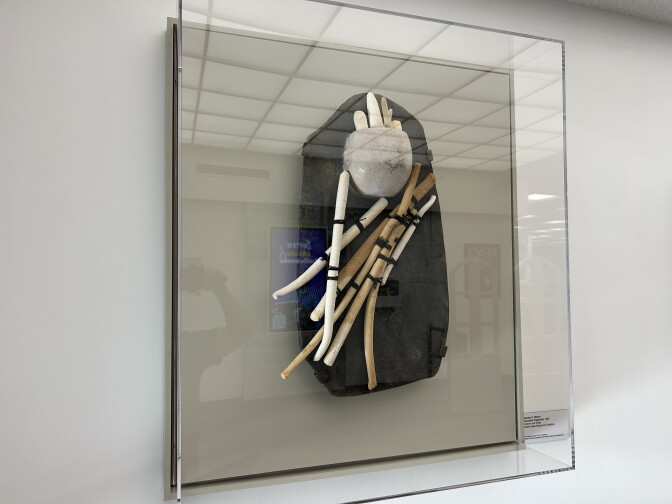 A sculpture made of ceramic with tubes and bowls is mounted on a board, and covered in a glass cover.
