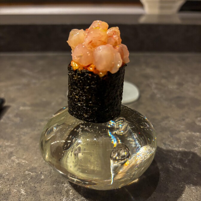 Close-up of a single piece of sushi presented on a decorative glass stand at a restaurant counter. The sushi features pink raw fish or seafood topped with what appears to be uni (sea urchin) or roe, wrapped in dark nori seaweed.