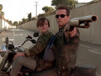 A young boy and a man wearing sunglasses and a leather jacket sit on a motorcycle, turned sideways in a flood-control channel. The man is pointing a rifle at something behind them while the boy looks at the man's face.