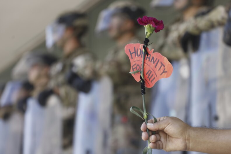 A hand holds a purple carnation flower with a paper attached to it that reads 'Humanity For All."