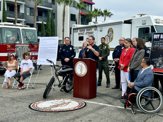A man speaks at an Orange County District Attorney podium beside an e-bike, with officials and vehicles from fire, sheriff and DA departments behind him.
