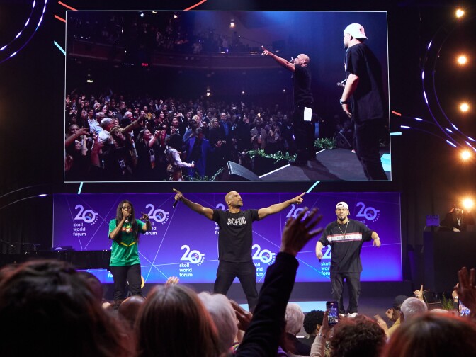 Three people stand and perform on an illuminated stage with the big screen above it. The screen shows camera perspective from the side, with the performers on the right and a large crowd on the left. The performer in the center, a bald Black man dressed in a black t-shirt and black pants, has his arms outstretched to the crowd.